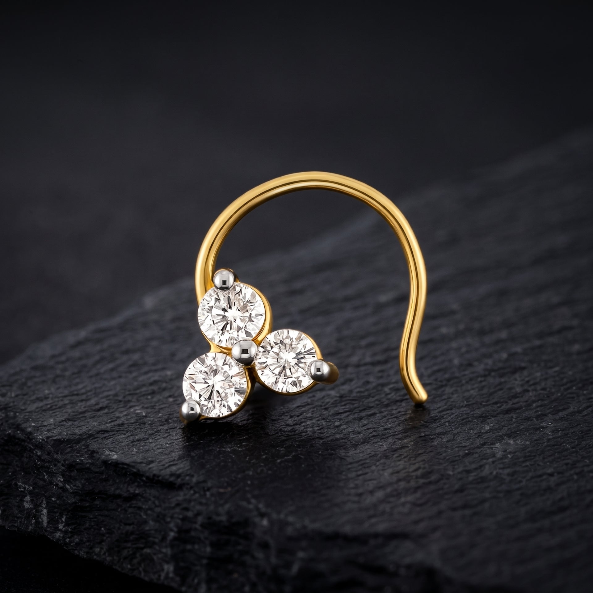 A gold crescent-shaped nose ring with three round clear gemstones, displayed on a dark textured slate surface.