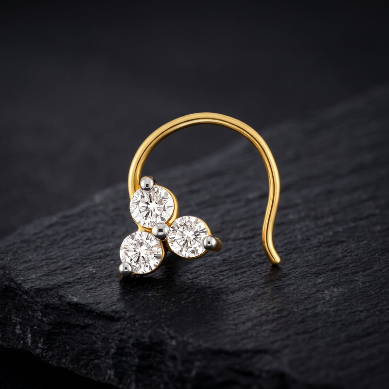 A gold crescent-shaped nose ring with three round clear gemstones, displayed on a dark textured slate surface.