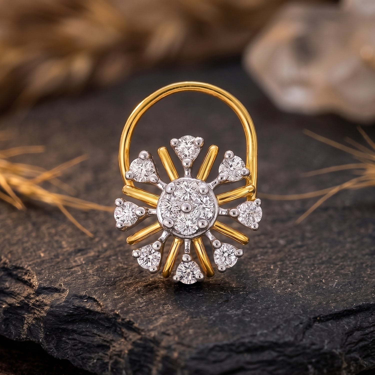 A gold and white gold sunburst ring with clear gemstones, displayed on a dark textured stone surface with dried grass and a blurred background.