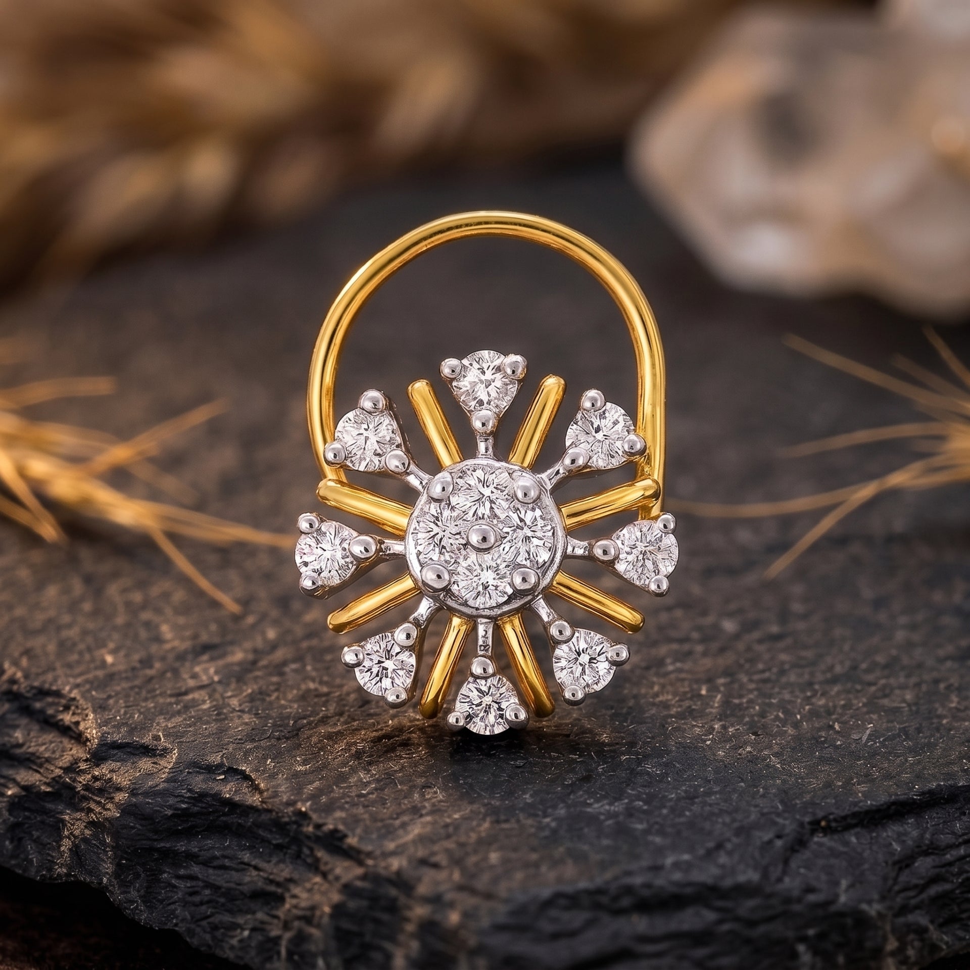 A gold and white gold sunburst ring with clear gemstones, displayed on a dark textured stone surface with dried grass and a blurred background.