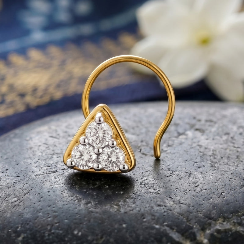 A gold triangular stud nose ring with a pyramid arrangement of clear gemstones, resting on a dark textured stone with a blurred white flower and blue background.