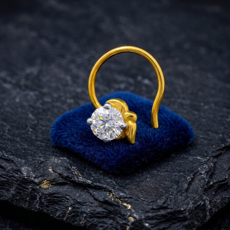 A gold nose ring with a round clear gemstone displayed on a small blue velvet cushion over a dark textured stone surface.