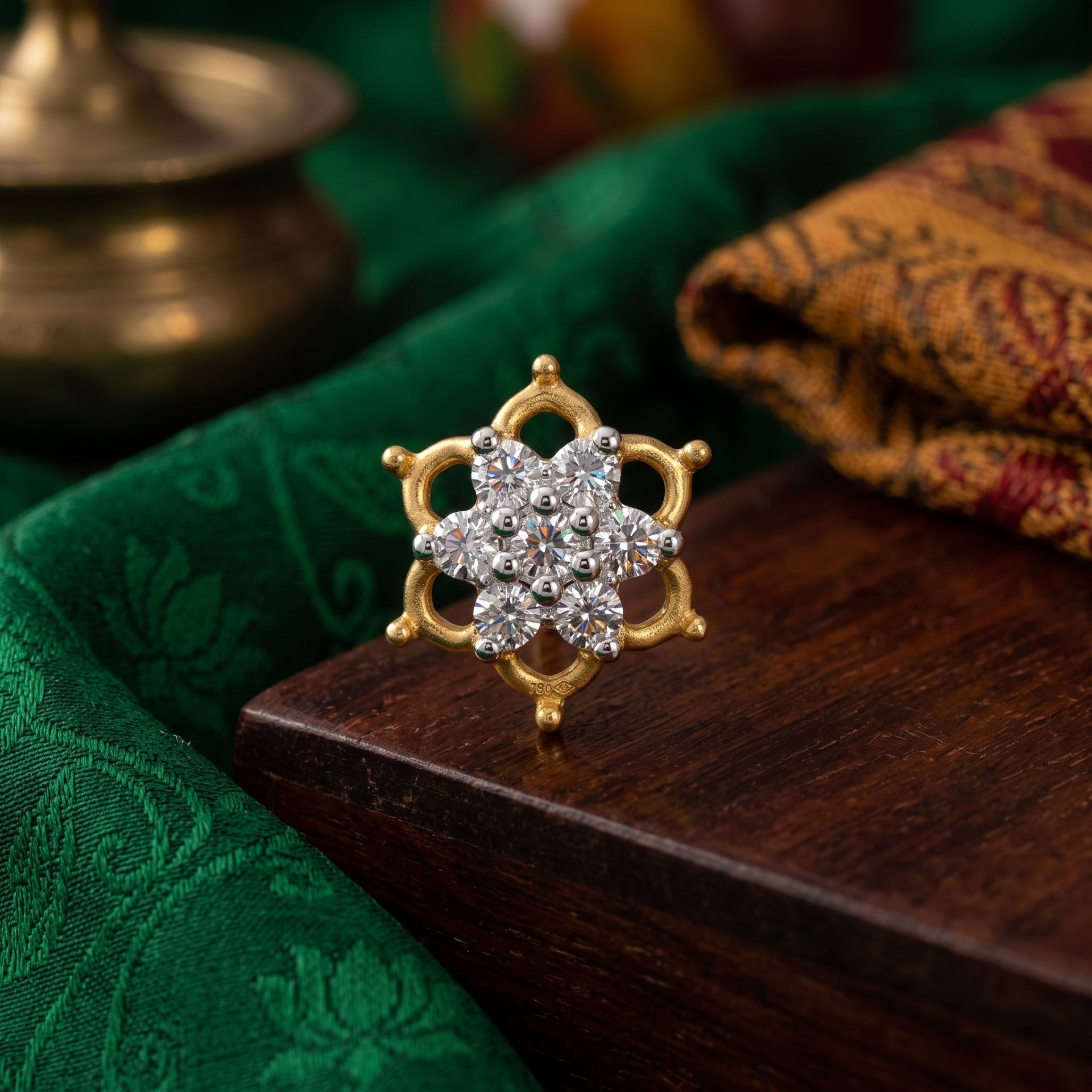 A gold ring with a floral cluster of clear stones, displayed on a dark wooden block with green and patterned fabric in the background.