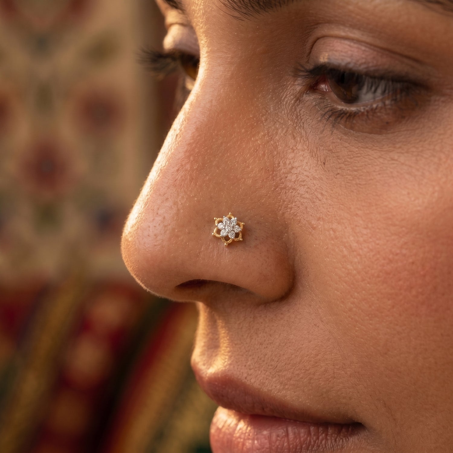 A close-up side profile of a woman wearing a small gold nose stud with a clear gemstone cluster on her nose, with a softly blurred patterned background.