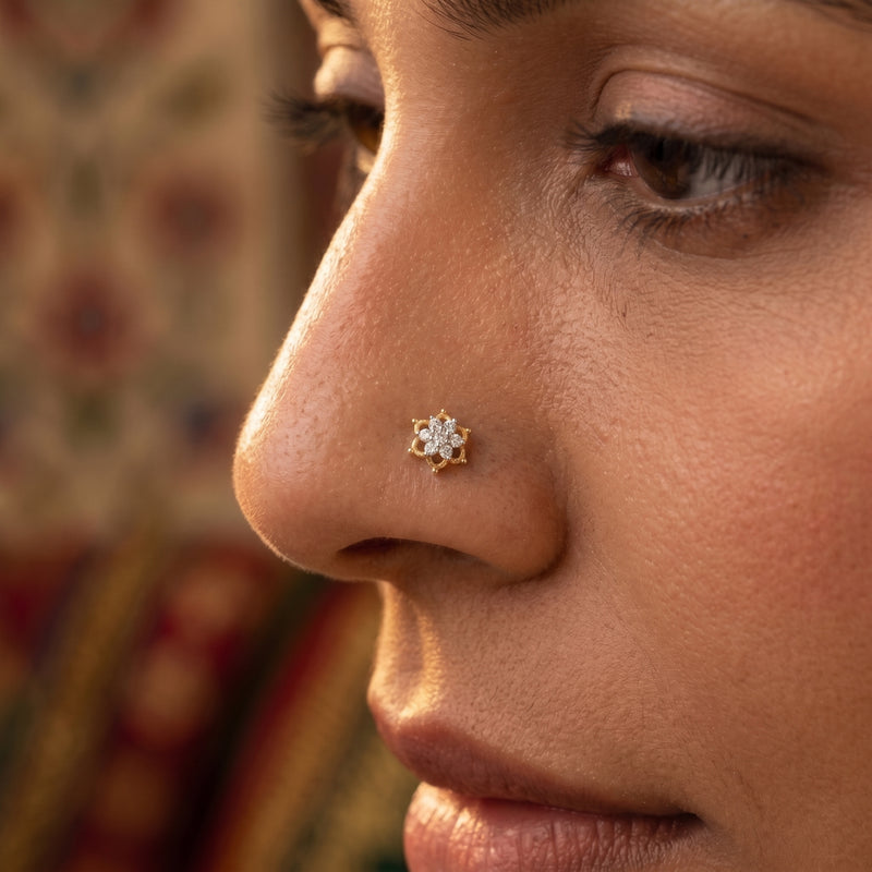 A close-up side profile of a woman wearing a small gold nose stud with a clear gemstone cluster on her nose, with a softly blurred patterned background.