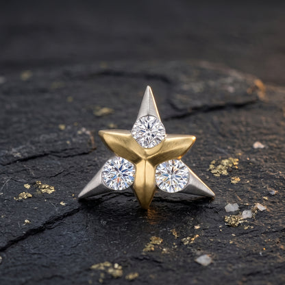 A star-shaped nose ring featuring three round-cut clear gemstones set in a mix of yellow gold and silver-toned metal, displayed on a dark textured surface with scattered gold flakes.