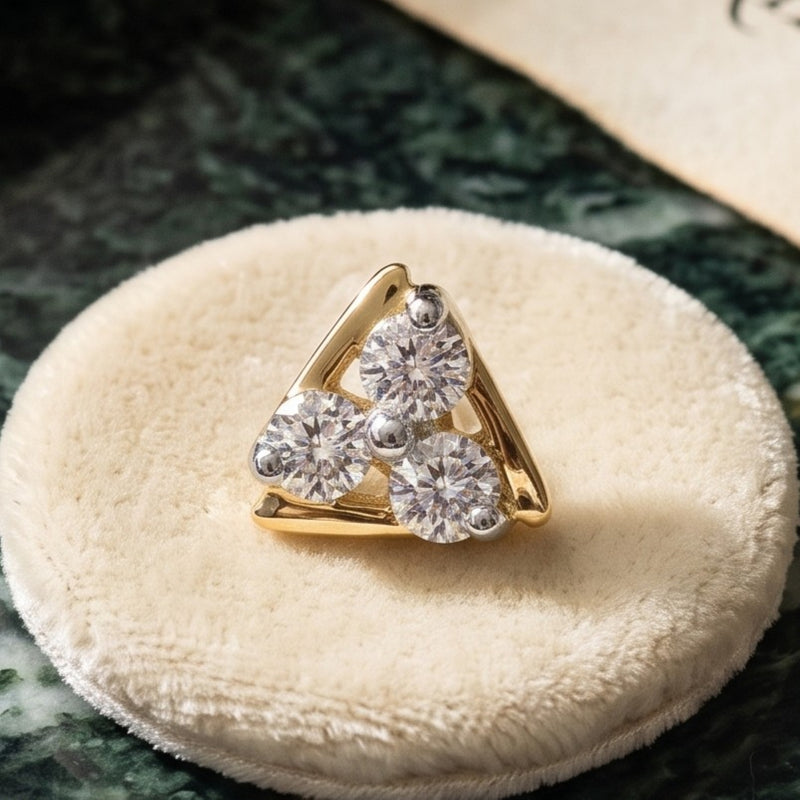 A gold triangular nose ring with three round clear gemstones, displayed on a round cream velvet ring cushion over a dark green marbled surface.