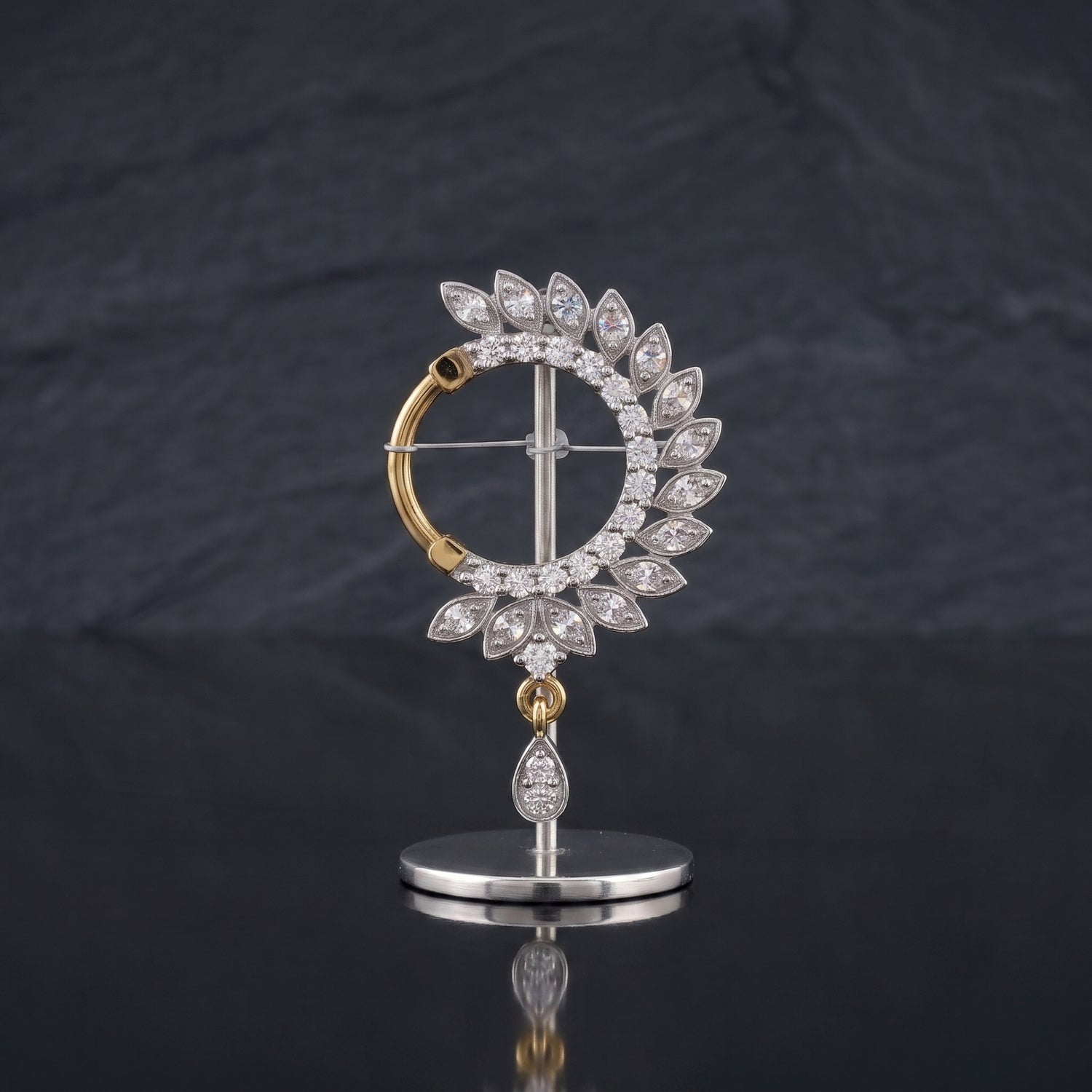 A gold and silver-tone nosepin featuring a circular halo of clear teardrop-cut stones, with a dangling teardrop stone on a polished stand, set against a dark slate background.