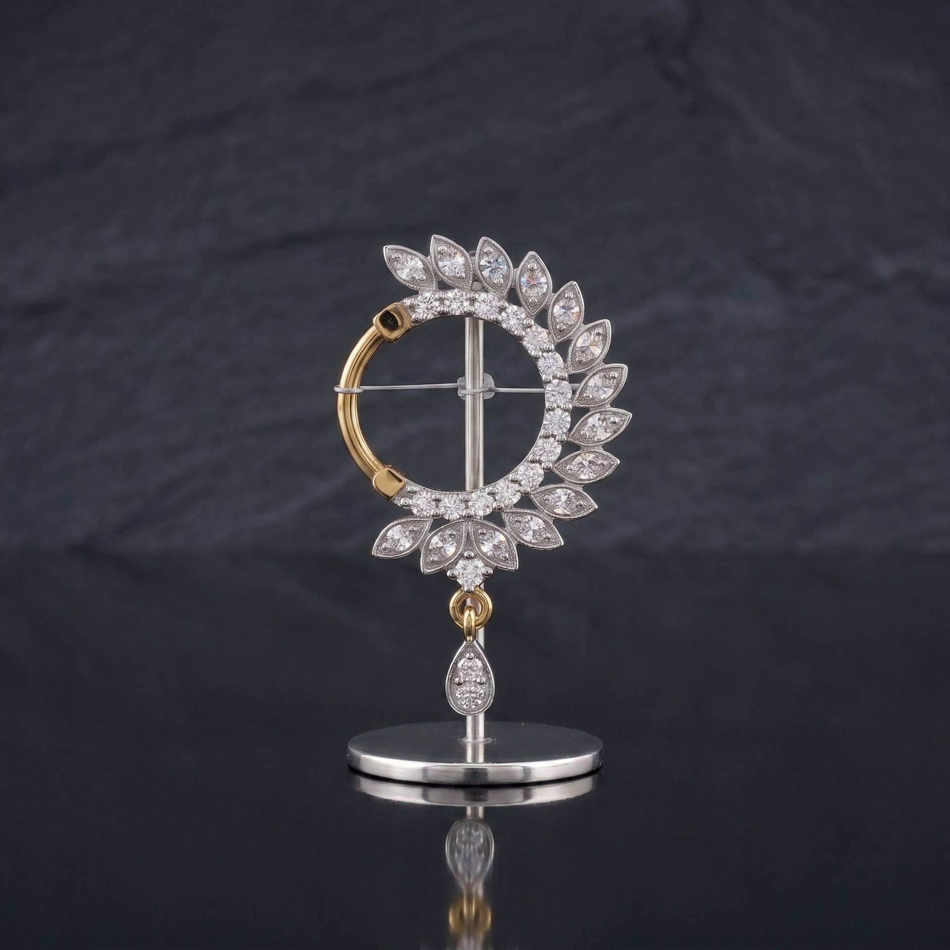 A gold and silver-tone nosepin featuring a circular halo of clear teardrop-cut stones, with a dangling teardrop stone on a polished stand, set against a dark slate background.