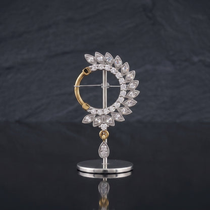 A gold and silver-tone nosepin featuring a circular halo of clear teardrop-cut stones, with a dangling teardrop stone on a polished stand, set against a dark slate background.