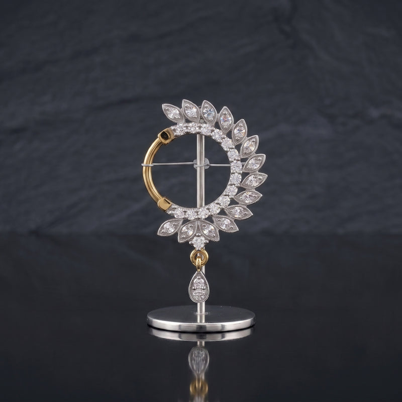 A gold and silver-tone nosepin featuring a circular halo of clear teardrop-cut stones, with a dangling teardrop stone on a polished stand, set against a dark slate background.