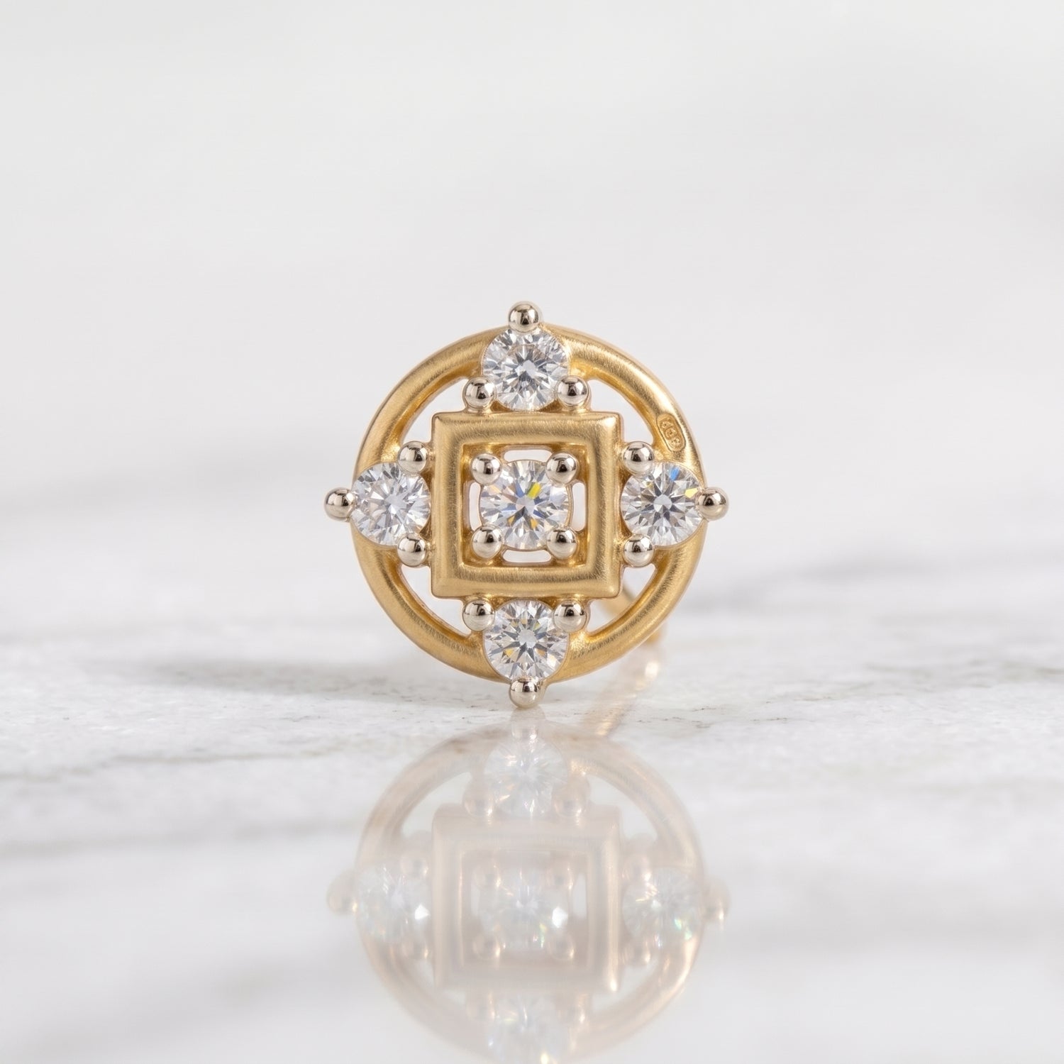 A yellow gold nose ring with a square and circular diamond setting, displayed on a light marble surface with a soft reflection.