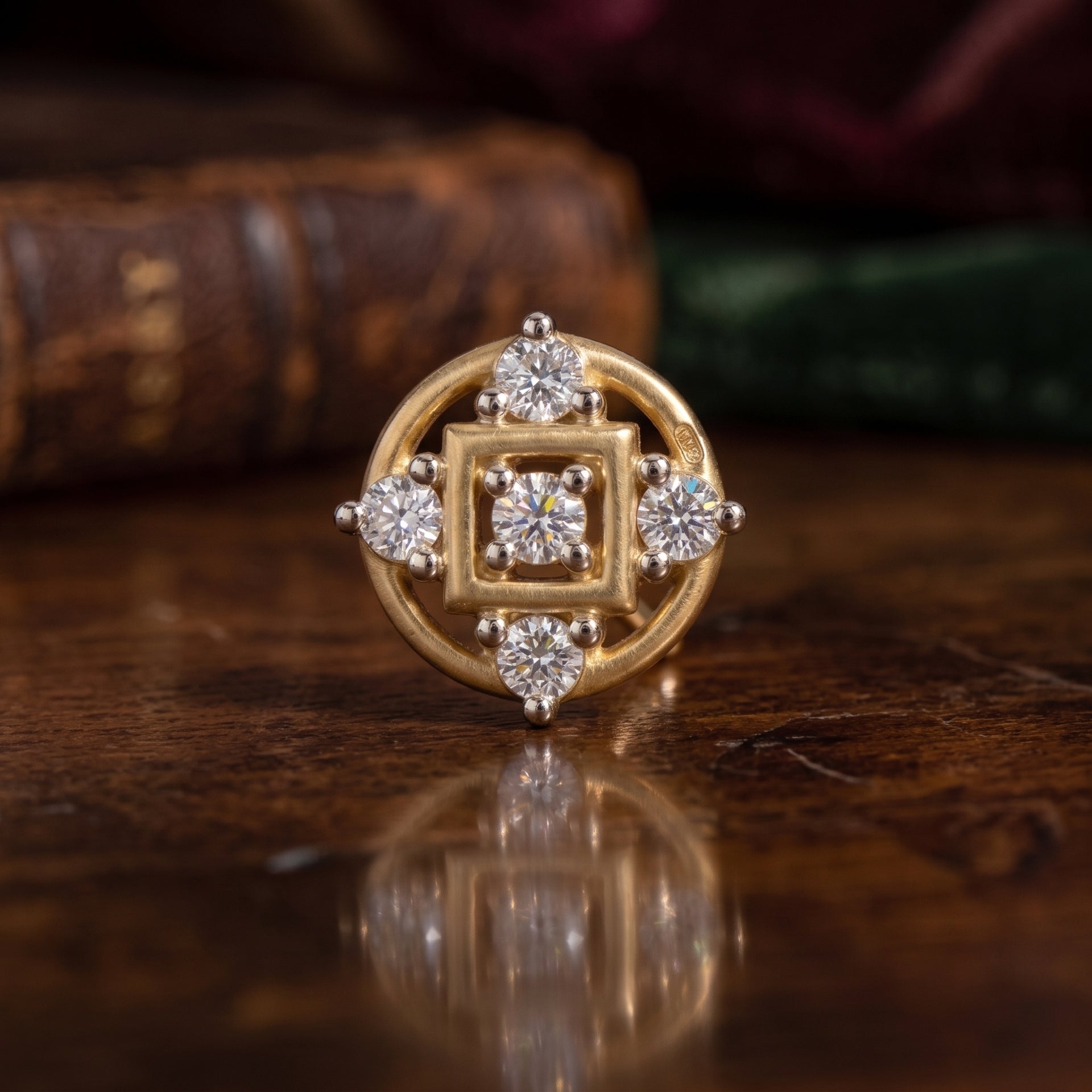 A gold nose ring with a square and circular geometric design, accented with four round-cut diamonds, displayed on a polished wooden surface with a blurred background.