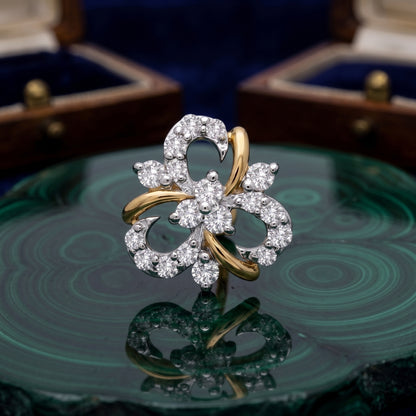 A floral-shaped ring with white gold or platinum pavé-set clear stones and curved yellow gold accents, displayed on a green malachite slab with blurred jewelry boxes in the background.