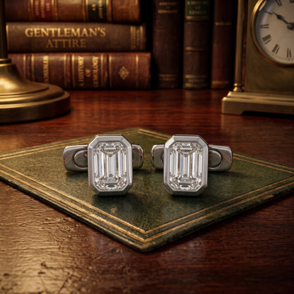A pair of silver-toned cufflinks with large rectangular baguette-cut clear stones, displayed on an old green book on a wooden desk with vintage books and a clock in the background.