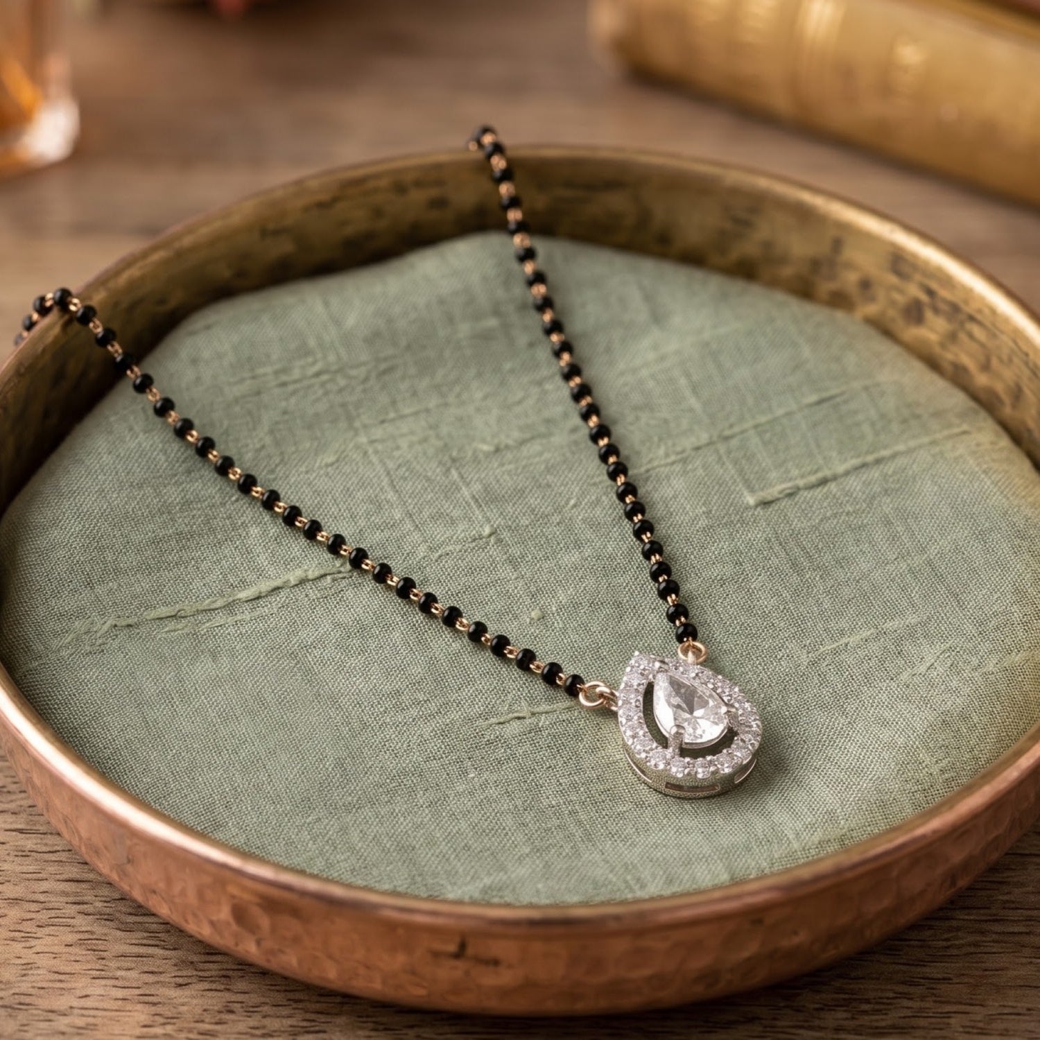 A black and gold beaded necklace with a silver-toned teardrop pendant set with clear stones, displayed on a textured sage green fabric cushion inside a rustic bronze-colored tray.