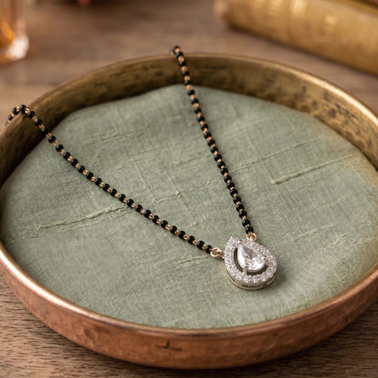 A black and gold beaded necklace with a silver-toned teardrop pendant set with clear stones, displayed on a textured sage green fabric cushion inside a rustic bronze-colored tray.