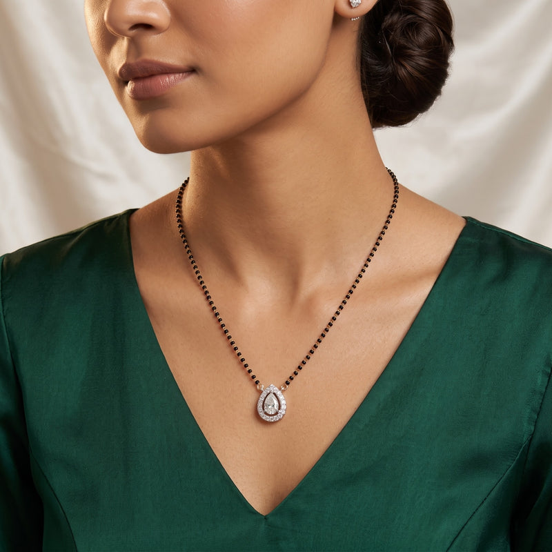 A close-up lifestyle shot of a model wearing a teardrop-shaped pendant necklace with a silver-toned halo and a black beaded chain, paired with a dark green satin top.
