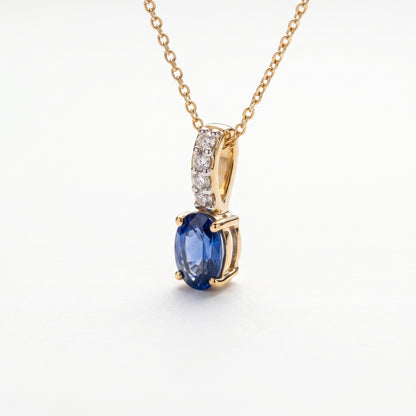A gold necklace with an oval blue sapphire pendant and a pavé-set diamond accent bar, on a wite background