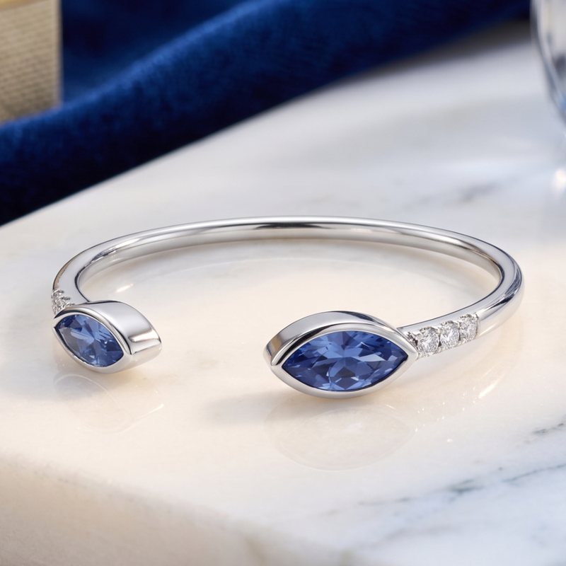 A polished silver cuff bracelet with two marquise-cut blue gemstones and a row of small clear stones on one side, displayed on a white marble surface.