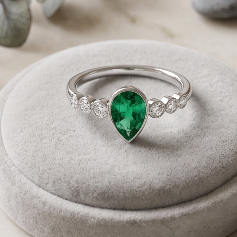 A silver ring featuring a large pear-cut green emerald center stone and smaller round clear stones along the band, displayed on a round light gray velvet ring stand.