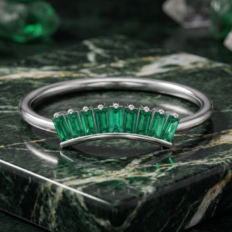 A silver bangle bracelet featuring a row of emerald-cut green gemstones, displayed on a polished green marble block.
