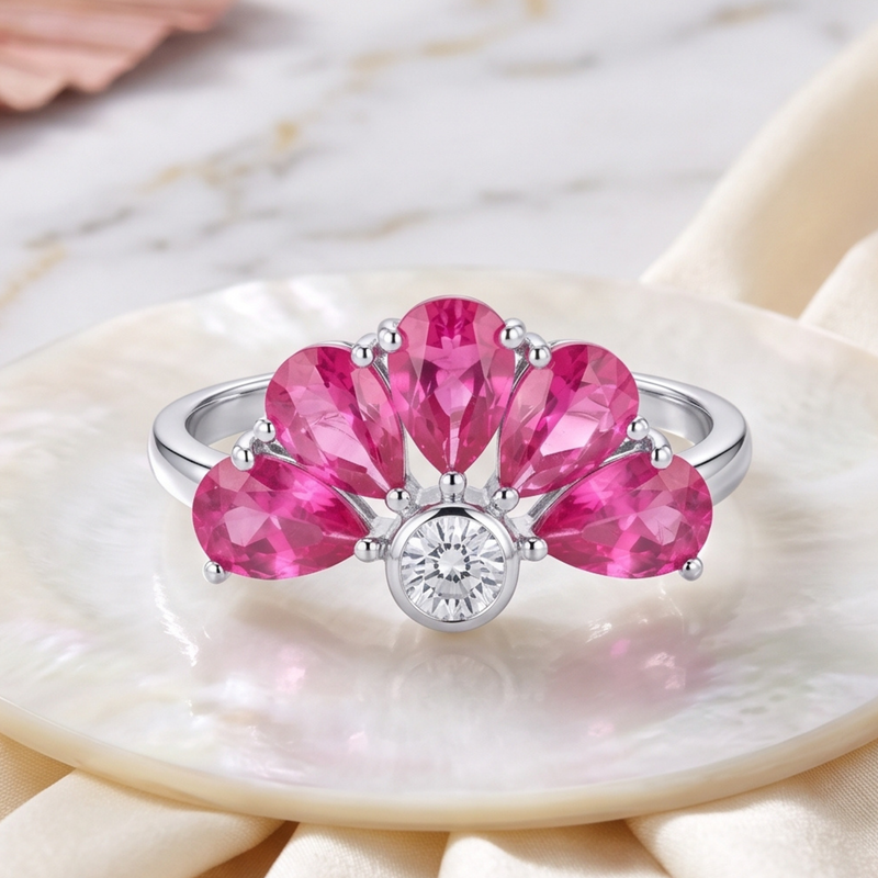 A silver ring featuring five bright pink marquise-cut gemstones arranged in a fan shape around a central round clear gemstone, displayed on a white shell dish over cream fabric.