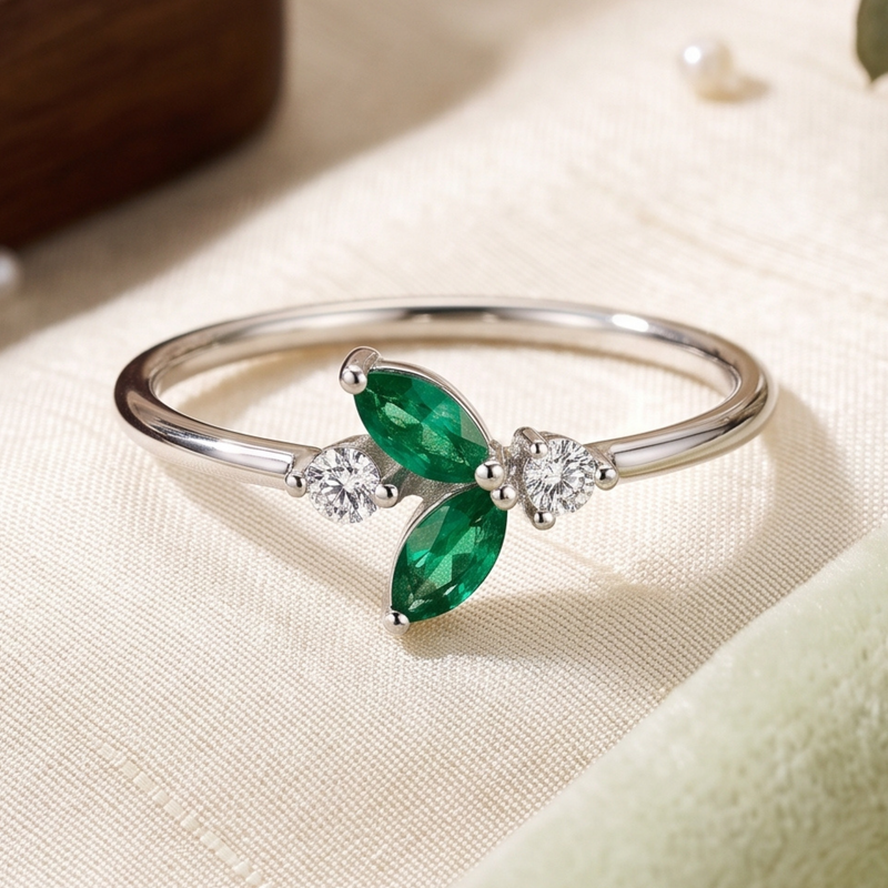 A silver bangle ring featuring two marquise-cut green emeralds and two small round clear stones, displayed on a textured cream fabric surface.
