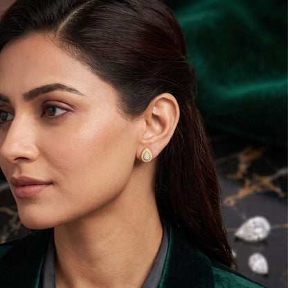 A close-up of a woman wearing a small teardrop-shaped gold earring with a clear stone, set against a dark green velvet background with decorative stones.