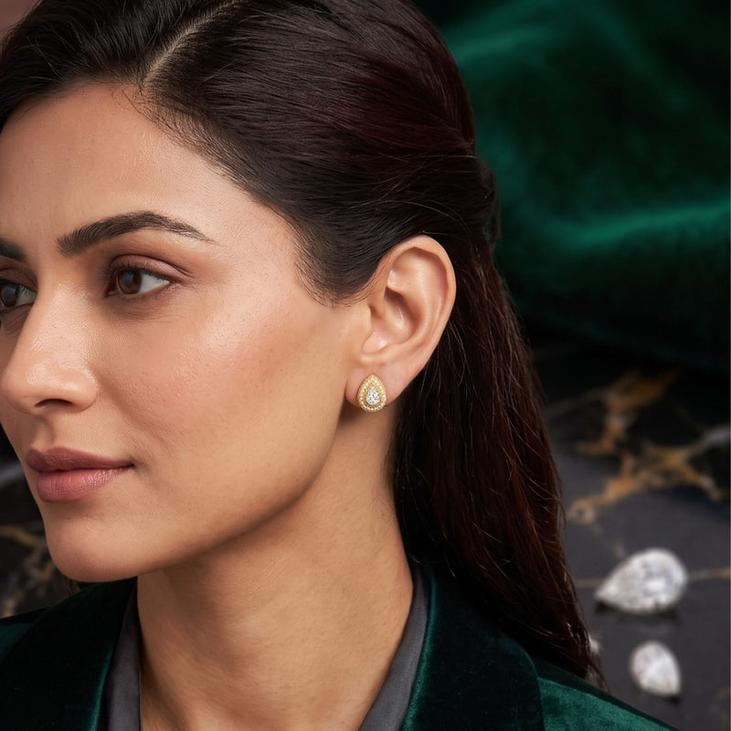 A close-up of a woman wearing a small teardrop-shaped gold earring with a clear stone, set against a dark green velvet background with decorative stones.