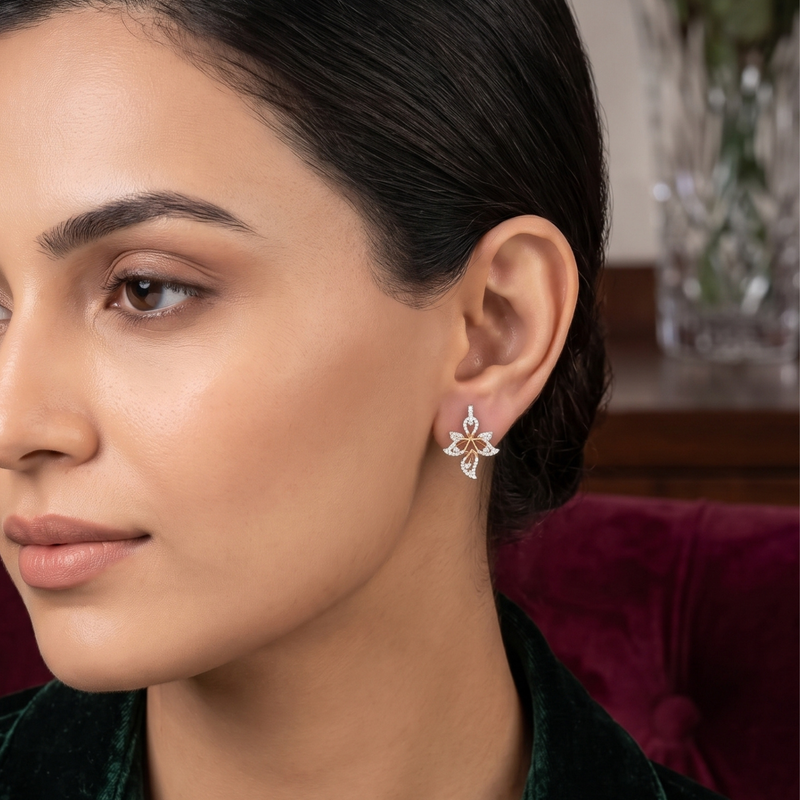 A close-up of a woman wearing a delicate silver-toned floral stud earring with small gold and white accents, set against a softly blurred indoor background.