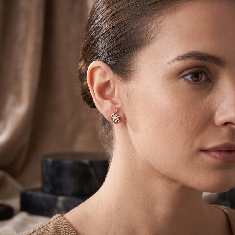 A close-up of a woman wearing a small floral stud earring with a silver-toned metal setting and clear stones, set against a warm brown draped background.
