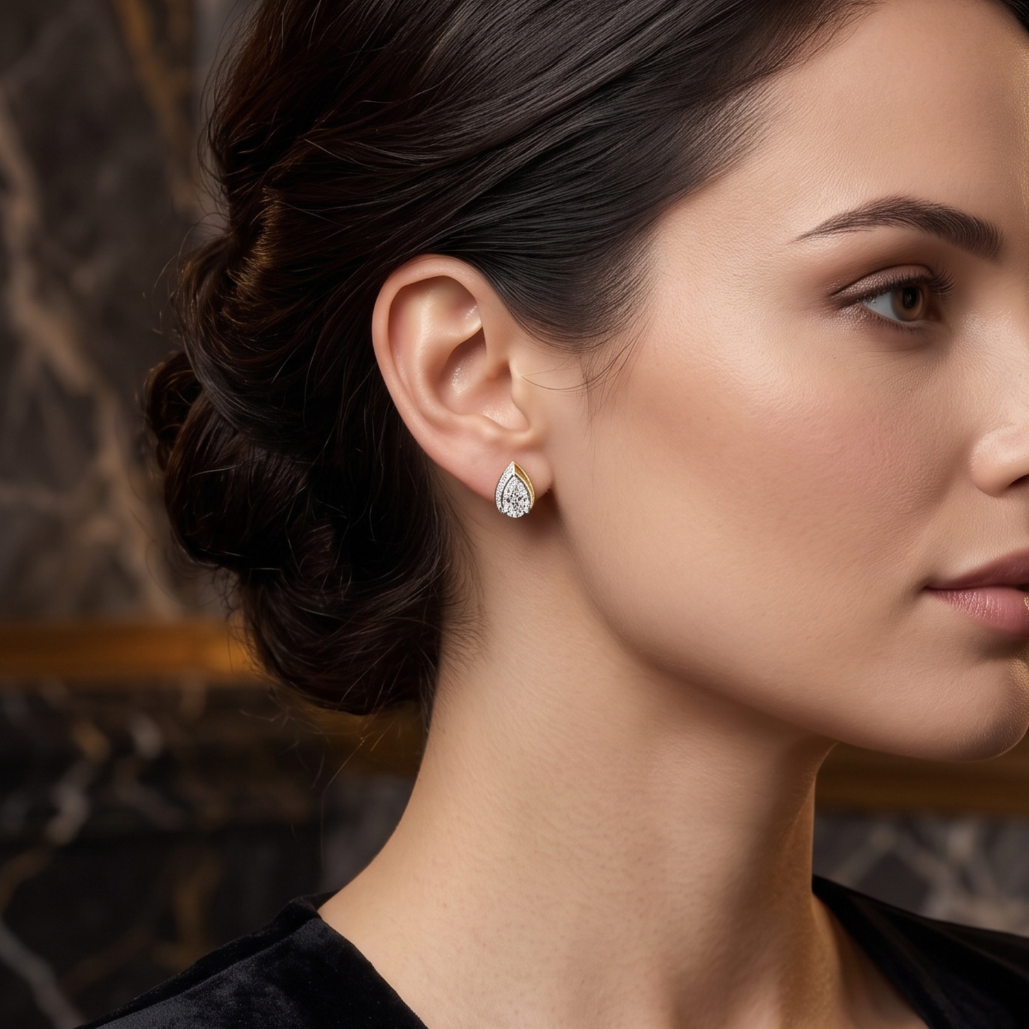 A close-up side profile of a woman wearing a small teardrop-shaped diamond stud earring in a silver setting, with a dark hair bun and a blurred dark marble background.