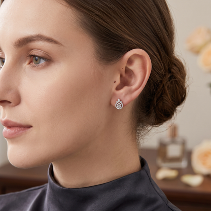 A close-up side profile of a woman wearing a small teardrop-shaped silver stud earring with clear gemstones, set against a softly blurred indoor background.