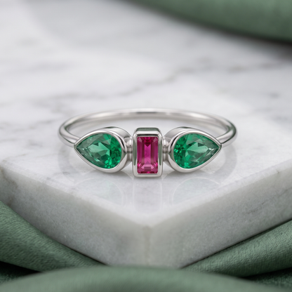 A silver ring featuring two green marquise-cut gemstones on either side of a central rectangular pink gemstone, displayed on a white marble block with soft green fabric in the background.