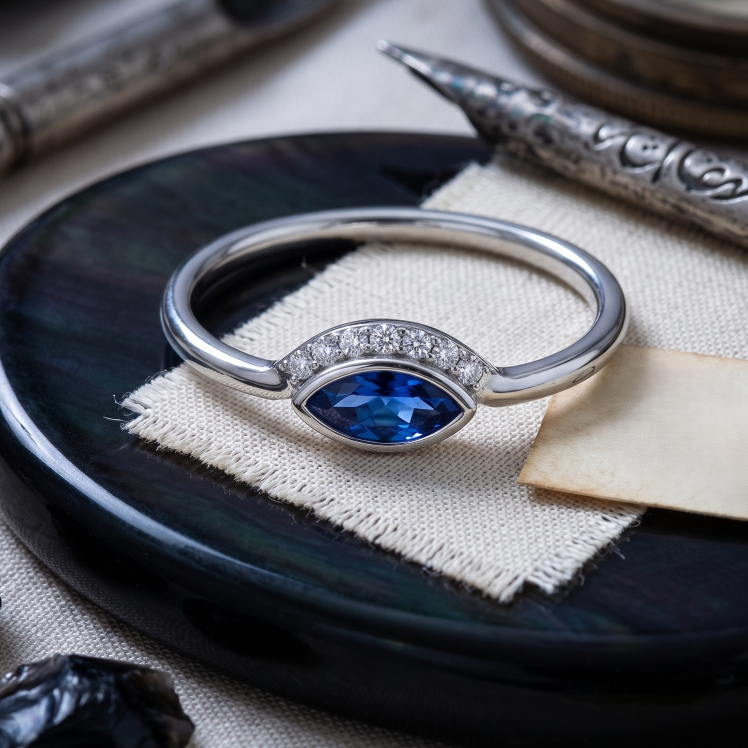A silver ring with a marquise-cut blue gemstone and a row of small clear stones, displayed on a textured cream fabric over a dark round tray.