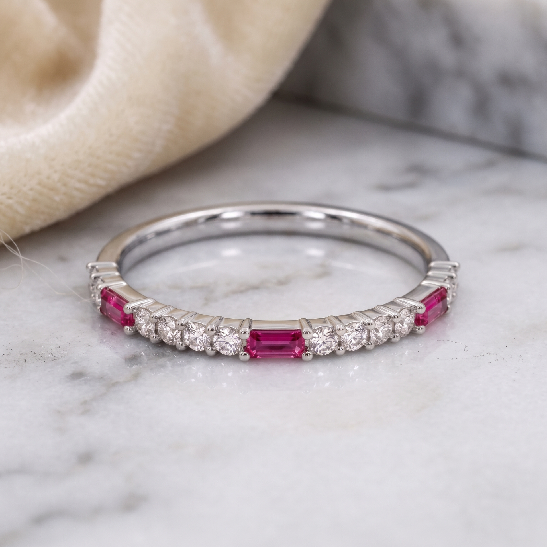 A silver band ring featuring a row of round clear stones and rectangular pink gemstones, resting on a white marble surface with a soft beige fabric in the background.