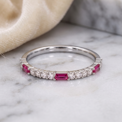 A silver band ring featuring a row of round clear stones and rectangular pink gemstones, resting on a white marble surface with a soft beige fabric in the background.