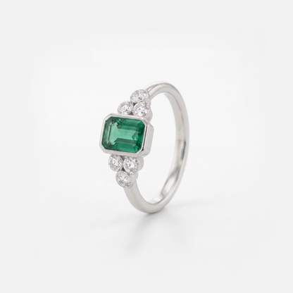 A silver ring featuring a rectangular green emerald-cut center stone and small clear round stones on either side of the band, photographed on a clean white background.