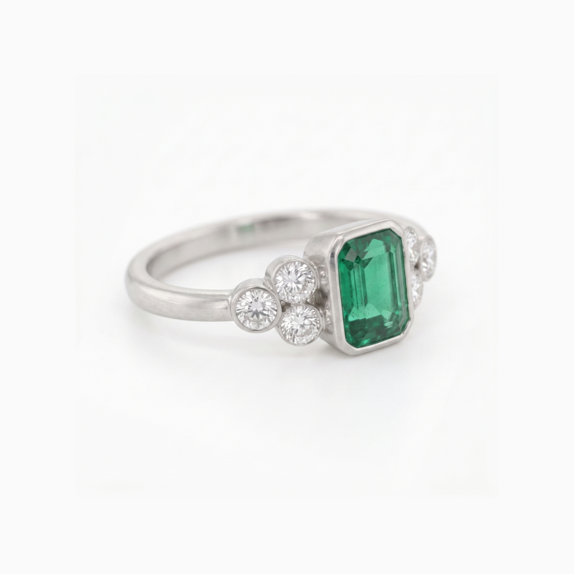 A silver ring featuring a rectangular emerald-cut green gemstone center, flanked by small round clear stones on each side, set against a plain white background.