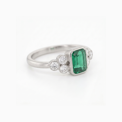 A silver ring featuring a rectangular emerald-cut green gemstone center, flanked by small round clear stones on each side, set against a plain white background.
