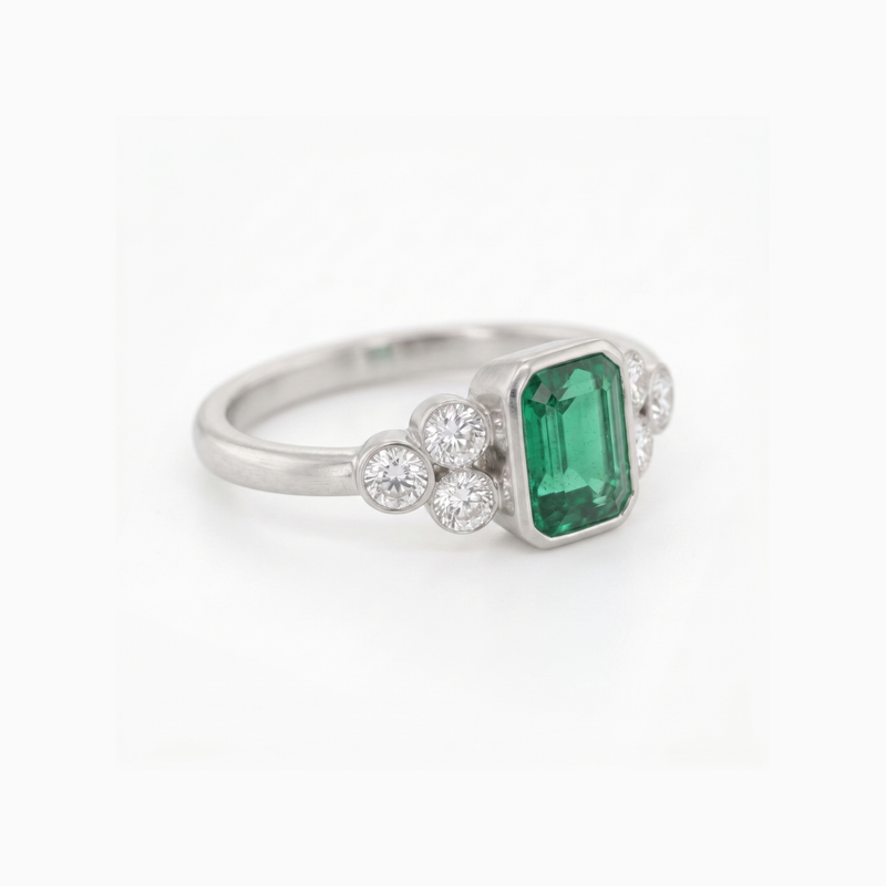 A silver ring featuring a rectangular emerald-cut green gemstone center, flanked by small round clear stones on each side, set against a plain white background.