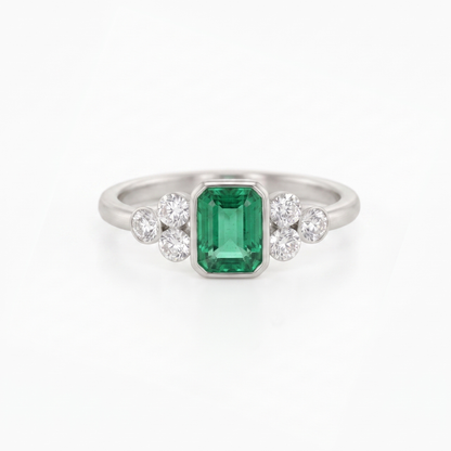 A silver ring featuring a central emerald-cut green gemstone flanked by two small clusters of round clear stones, set against a plain white background.