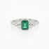 A silver ring featuring a central emerald-cut green gemstone flanked by two small clusters of round clear stones, set against a plain white background.