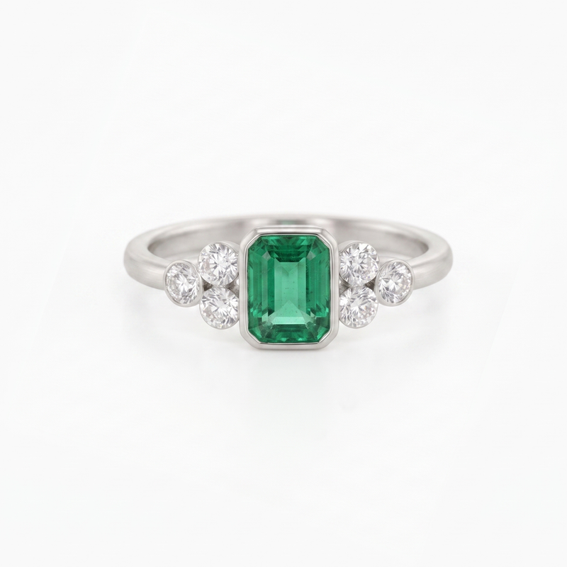 A silver ring featuring a central emerald-cut green gemstone flanked by two small clusters of round clear stones, set against a plain white background.